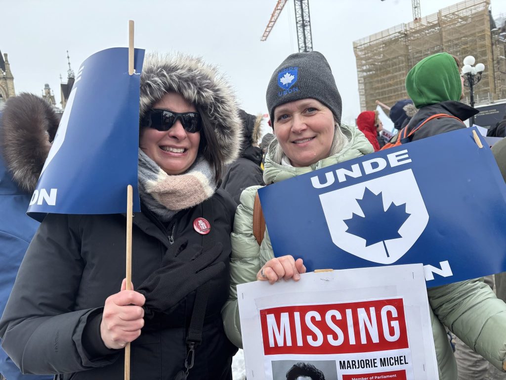Two UNDE members outdoors in winter, one wearing a UNDE toque, holding a UNDE placard and an awareness sign